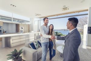 Real estate agent with couple in luxury home. They are shaking hands. There is a water view, kitchen and living room in the background. Couple are casually dressed. They are laughing. Agent is dressed in a suit and smiling. Wide angle.